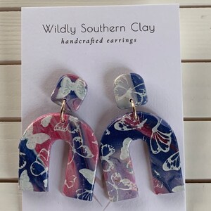 Könnte beinhalten: Handgefertigte Ohrringe mit einem marmorierten Design mit weißen Schmetterlingsmustern. Die Ohrringe haben ein blaues, rosa und lila Farbschema. Der Text "Wildly Southern Clay" und "handcrafted earrings" ist auf der Karte zu sehen.