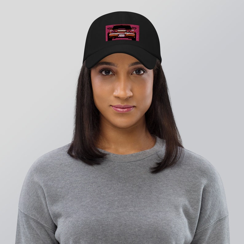 May include: Black baseball cap featuring a stylized image of a classic car in shades of pink and red. The car graphic is on the front of the cap, set against a pink background. The cap is worn by a person.