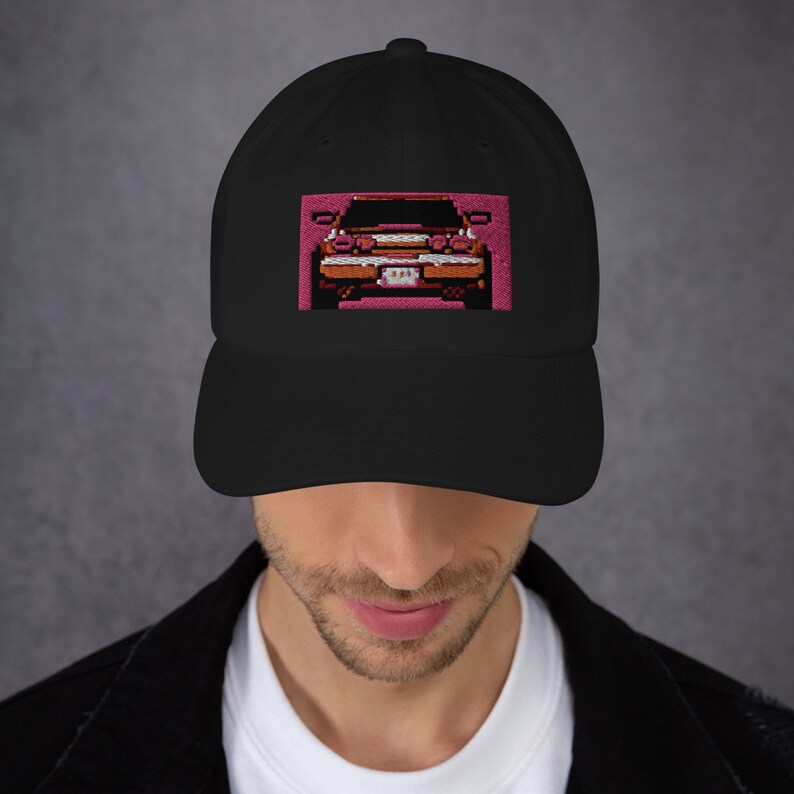 May include: Black baseball cap with a pixelated car design embroidered on the front. The car is depicted in shades of brown, orange, and white against a pink background. The cap has a curved brim and a classic six-panel design.