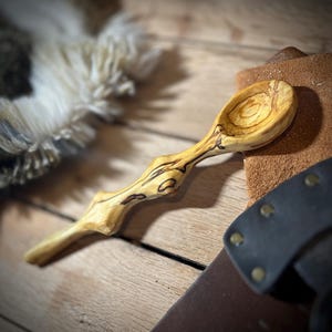 May include: Hand-carved wooden spoon with a light brown and dark brown grain pattern. The spoon has a curved handle and a rounded bowl. The wood grain is visible throughout. Likely for kitchen use.