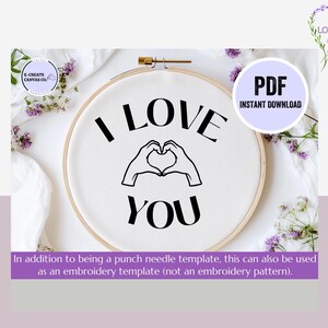 May include: A black and white embroidery template with the text "I LOVE YOU" and a heart shape formed by two hands. The template is displayed on a white fabric background in a round embroidery hoop.