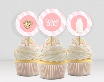 May include: Three cupcakes with white icing and colourful sprinkles. Each cupcake has a pink and white striped topper. One topper has a cookie, another says "sweet one", and the last has a milk bottle.