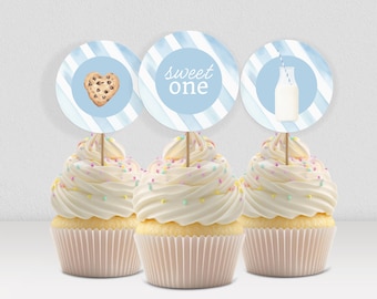May include: Three cupcakes with white icing and colourful sprinkles. Each cupcake has a round topper with a blue and white striped background. The toppers feature a cookie, the text "sweet one", and a milk bottle.
