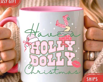 May include: White ceramic mug with a pink interior and handle. The mug features the text "Have a Holly Dolly Christmas" in pink and green, along with illustrations of a Santa hat, cowboy boots, and a kiss.