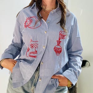 May include: A blue and white striped button-down shirt with red embroidered designs. The designs include a face, a vase with flowers, and a heart.