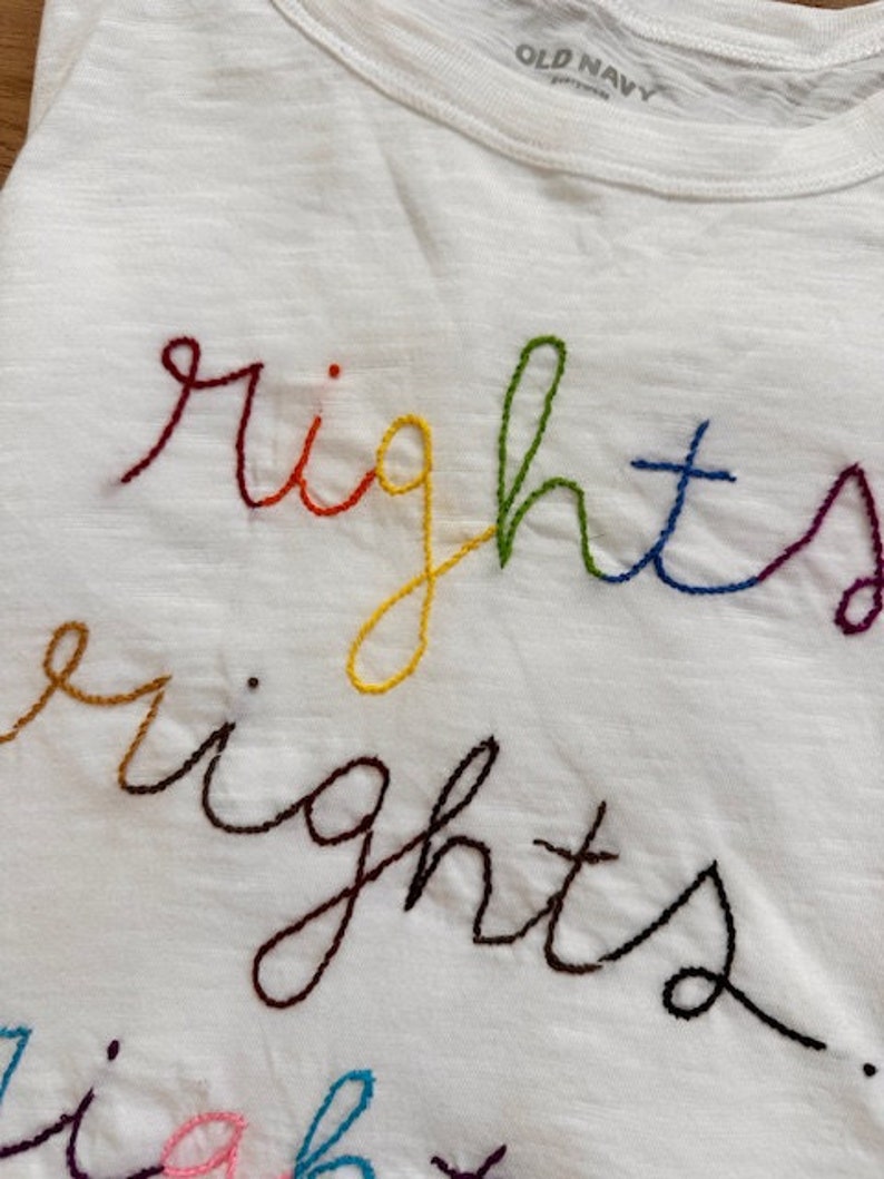 May include: White t-shirt with the word "rights" embroidered in rainbow colours.