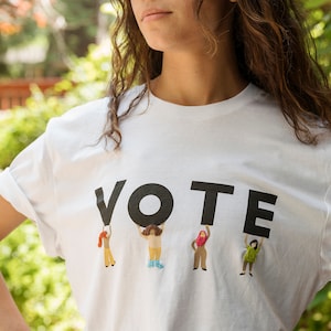 May include: A white t-shirt with the word "VOTE" printed in black letters.  Five small figures are holding up the letters.  The figures are all different colors and have different hairstyles.