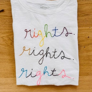 May include: White t-shirt with the word "rights" embroidered three times in rainbow colours. The shirt has a tag that says "Old Navy" on the neck.