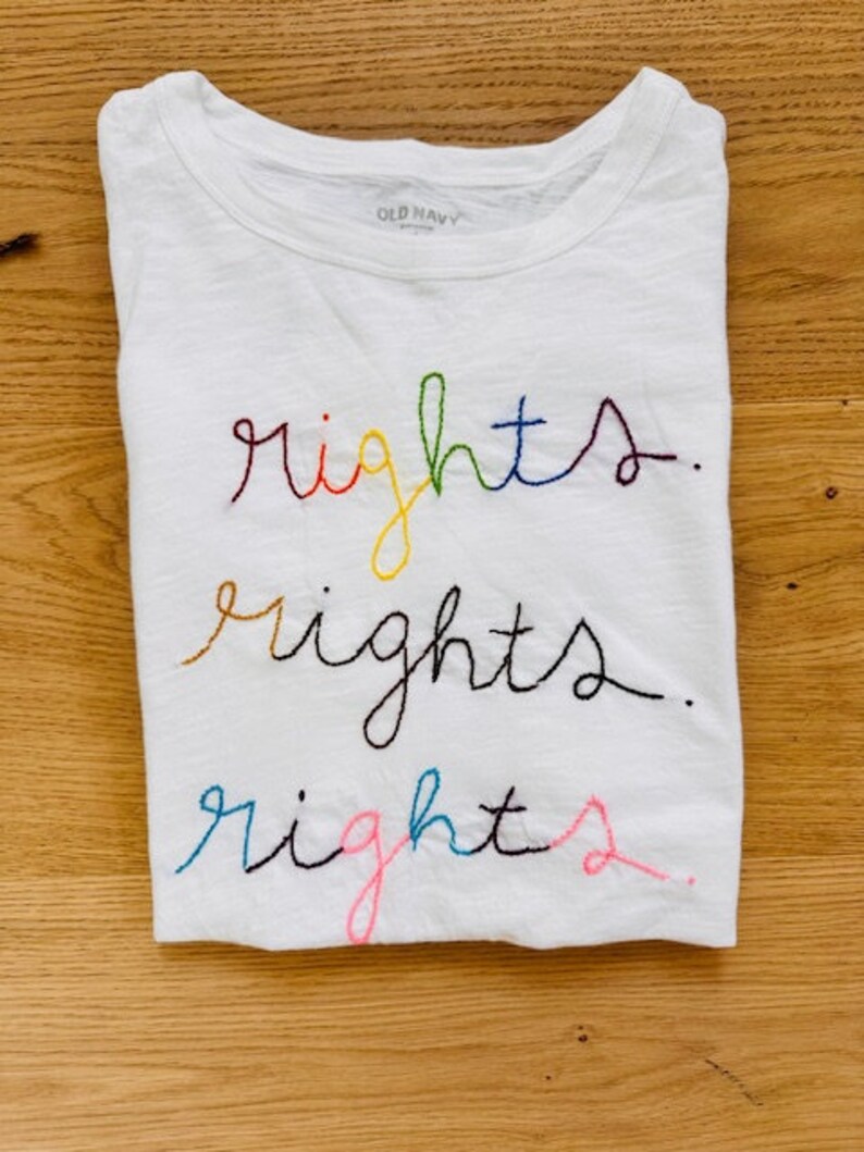 May include: White t-shirt with the word "rights" embroidered three times in rainbow colours. The shirt has a tag that says "Old Navy" on the neck.