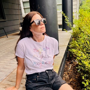 May include: A woman wearing a white t-shirt with the words "rights" written in rainbow colours. She is wearing black denim shorts and sunglasses.