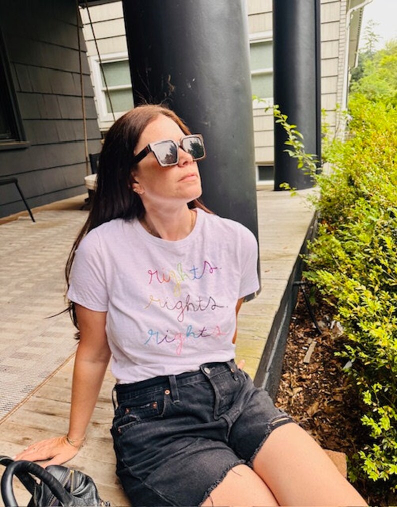 May include: A woman wearing a white t-shirt with the words "rights" written in rainbow colours. She is wearing black denim shorts and sunglasses.