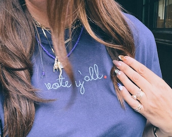 May include: A navy blue short-sleeved t-shirt featuring the embroidered text "vote y'all." in white cursive script, with a small red embroidered dot at the end of the sentence. The shirt has a crew neckline and a simple, solid-coloured fabric design.
