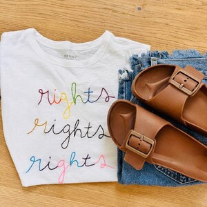 May include: White t-shirt with the word "rights" embroidered in rainbow colours. The shirt is paired with blue denim jeans and brown leather sandals.
