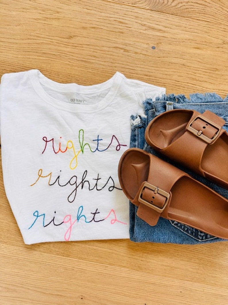 May include: White t-shirt with the word "rights" embroidered in rainbow colours. The shirt is paired with blue denim jeans and brown leather sandals.