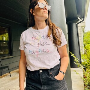 May include: A white t-shirt with the word "rights" embroidered in rainbow colours. The person wearing the shirt is standing in front of a black column.