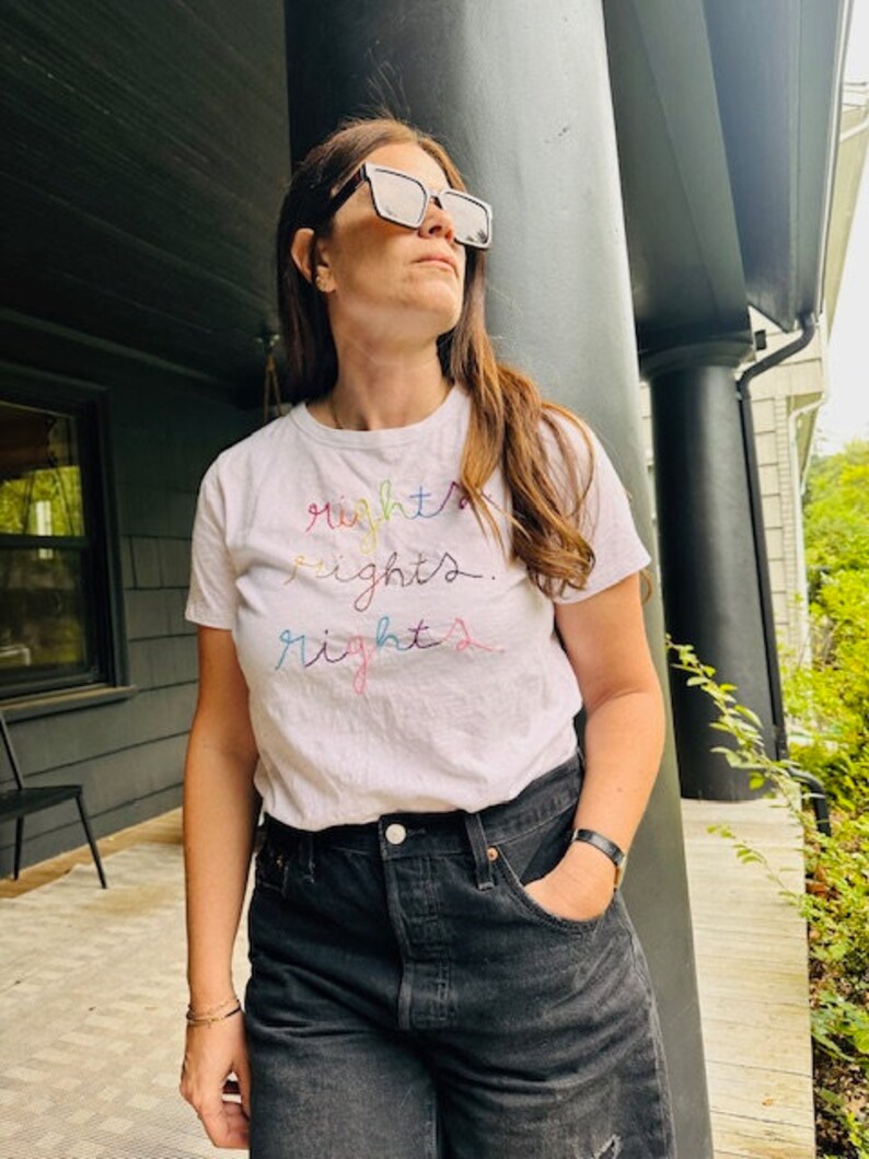 May include: A white t-shirt with the word "rights" embroidered in rainbow colours. The person wearing the shirt is standing in front of a black column.