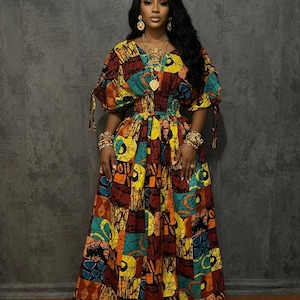 May include: A vibrant, floor-length dress with a colorful patchwork design. The dress features a V-neckline, short sleeves with tie details, and a cinched waist. Gold jewelry complements the dress.