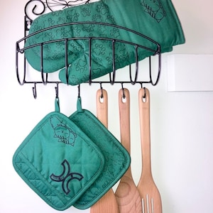 May include: A black metal rack with hooks holds green oven mitts and wooden kitchen utensils. The oven mitts have a paw print pattern and a cat design. The utensils include a wooden spoon and a slotted spatula.