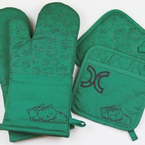 May include: A green kitchen set featuring an oven mitt and two pot holders. The oven mitt has a paw print pattern and a sleeping cat design. The pot holders also have the paw print pattern, a sleeping cat design, and the text "XC KITCHEN".