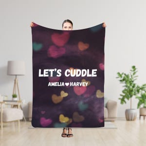 Puede incluir: Una manta de forro polar morada y negra con un fondo borroso de corazones. El texto "Let's Cuddle Amelia ♥ Harvey" está impreso en blanco en la manta.