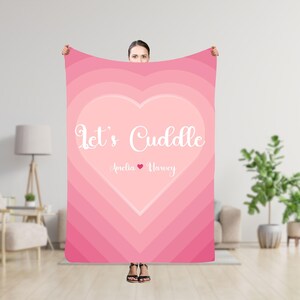 Puede incluir: Manta rosa y blanca con un gran diseño de corazón. El corazón tiene el texto "Let's Cuddle" y los nombres "Amelia" y "Harvey" con un pequeño símbolo de corazón entre ellos.