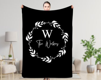 Könnte beinhalten: Schwarze Decke mit einem weißen floralen Kranzdesign. Der Kranz umgibt den Buchstaben "W" und den Text "The Walkers" in einer Schreibschrift. Die Decke wird von einer Person gehalten.