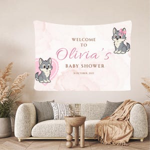 May include: A baby shower banner with the text "Welcome to Olivia's Baby Shower" in pink and brown. The banner features cartoon wolf illustrations and a pink watercolor background. The date of the baby shower is October 31, 2025.