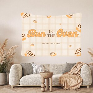 Peut inclure: Une tapisserie rectangulaire avec l'inscription "Bun in the Oven" en orange, et la date "03, JANVIER 2026". Le fond est un motif à carreaux beige clair avec des illustrations de petits pains à la cannelle. La tapisserie est affichée au-dessus d'un canapé.