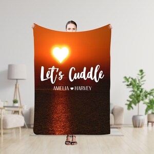Puede incluir: Una manta rectangular grande con una escena de puesta de sol y un sol en forma de corazón. Las palabras "Let's Cuddle" están escritas en blanco, con "AMELIA ♥ HARVEY" debajo. La manta es sostenida por una persona.