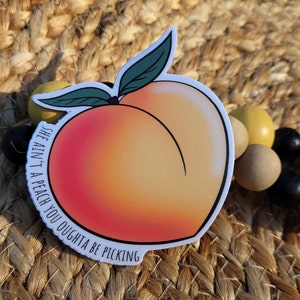 Puede incluir: Adhesivo con una ilustración de melocotón en tonos naranja y rosa. El adhesivo tiene el texto "SHE AIN'T A PEACH YOU OUGHTA BE PICKING". El melocotón tiene hojas verdes y un contorno negro.