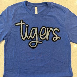 May include: Blue t-shirt with the word "tigers" in a sequined design. The shirt has a layered hem and a crew neck. The word "tigers" is outlined in black.