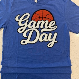 May include: A blue t-shirt with the words "Game Day" in white script, with a basketball graphic. The basketball is orange with black lines. The shirt is a crew neck and has a layered design.