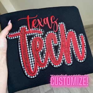 May include: Black sweatshirt with the words "Texas Tech" in red script and a red glitter outline. The letters are over a black and white checkered pattern. The word "CUSTOMIZE!" is in pink at the bottom.