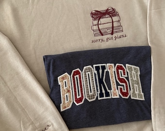 Könnte beinhalten: Beiges Sweatshirt mit einer Grafik von einem Bücherstapel und dem Text "sorry, got plans". Ein marineblaues Sweatshirt mit dem Wort "BOOKISH" in einem Patchwork-Design ist darüber gelegt. Der Ärmel des beigen Sweatshirts hat den Text "book lover".