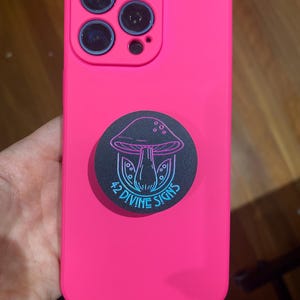 May include: A pink phone case with a black pop socket featuring a mushroom with the text "42 Divine Signs" in blue and pink.