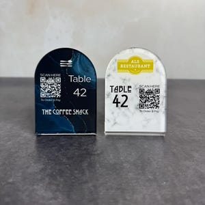May include: Two arched table signs. One is navy blue with a marble design, the text "Table 42", "The Coffee Shack", and a QR code. The other is white with a marble design, the text "Table 42", "ALS Restaurant", and a QR code.