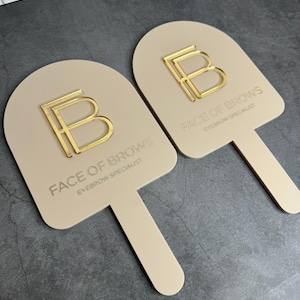 May include: Two beige, arch-shaped handheld signs with a handle. Each sign features a raised, gold-coloured letter B logo and the text FACE OF BROWS EYEBROW SPECIALIST printed in a gold-coloured font on the smooth, matte surface of the sign.