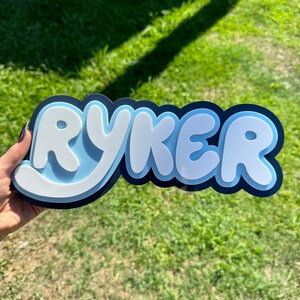 May include: A blue and white personalised name sign with the name "Ryker" in bubble letters.