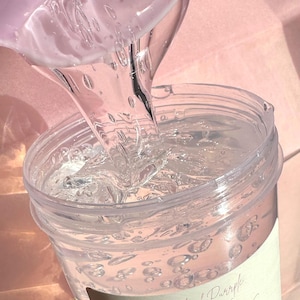Peut inclure: Gros plan sur du gloss transparent versé dans un récipient en plastique transparent. Le gloss a une texture mousseuse et porte l'étiquette "Pinkish Purple Collections LIP GLOSS" sur une étiquette crème. Le fond est rose pâle.