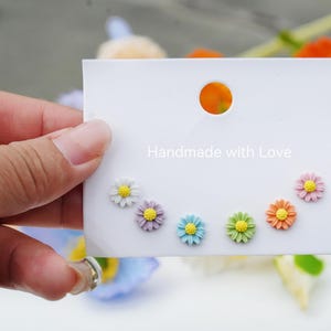 Op de afbeelding: Een witte kaart met de tekst "Handmade with Love" en zes kleurrijke madeliefjes oorbellen die erop worden getoond. De oorbellen zijn in tinten wit, lichtroze, lichtblauw, lichtgroen en oranje.