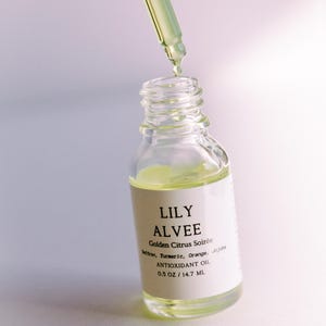 May include: A clear glass bottle with a dropper filled with a yellow liquid. The bottle is labeled "LILY ALVEE Golden Citrus Soirée Saffron, Turmeric, Orange, Jojoba ANTIOXIDANT OIL 0.5 OZ/14.7 ML."