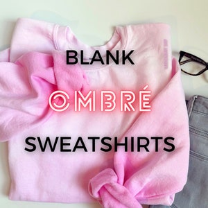 May include: A light pink ombre sweatshirt with the words "BLANK OMBRÉ SWEATSHIRTS" in black and red neon lettering. The sweatshirt is folded, with a pair of glasses and gray jeans in the background.