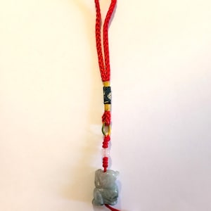 May include: A jade bear charm with a red braided cord and decorative beads. The bear is a light green color and has a detailed carving. The cord has a gold and black accent. The charm is designed to be hung.