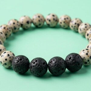 Peut inclure: Un bracelet élastique composé de perles rondes, alternant des perles de jaspe dalmatien couleur crème tachetées de brun foncé et quatre perles en pierre de lave noire poreuse et texturée, disposées en une boucle continue unique.