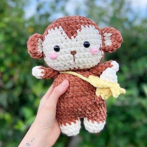 Könnte beinhalten: Handgehäkelte Affenpuppe in Braun und Weiß, die eine gelbe Banane hält. Der Affe hat schwarze Augen und rosa Wangen. Die Puppe ist weich und kuschelig, geeignet als Kinderspielzeug oder Dekoration für das Kinderzimmer.