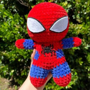 Könnte beinhalten: Handgemachte Spider-Man-Puppe aus Plüsch. Die Puppe hat einen roten Kopf und Körper mit blauen Akzenten an Armen und Beinen. Sie hat weiße Augen und ein schwarzes Spinnenemblem auf der Brust. Das Spielzeug ist weich und strukturiert.
