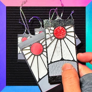 May include: Rectangular ornaments with a sun design. Each ornament features a red sun in the center, surrounded by white and gray geometric shapes, and outlined in black glitter. They have silver hanging strings.
