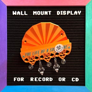 May include: Orange wall mount display for records or CDs. The display features a fan-shaped design with the text "THE LIFE OF A SH*RL" and decorative hanging crystals. The display is set against a black background.