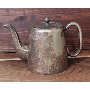 Op de afbeelding: Een vintage zilveren theepot met een gebogen tuit en een ronde handgreep. De theepot heeft een deksel met een kleine knop en vertoont tekenen van ouderdom en gebruik. Het zilveren oppervlak heeft een aangeslagen uiterlijk.