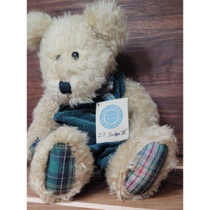 May include: A plush teddy bear with cream-colored fur, wearing a teal corduroy outfit and plaid accents on its paws. The bear has a black nose and a tag that reads "J.T. Edgar III". The bear is sitting on a wooden surface.
